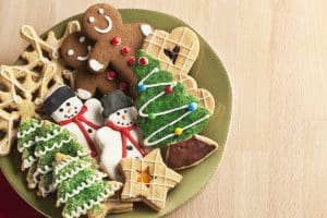 A plate filled with Christmas cookies decorated with frosting and icing for winter holiday celebrations. The baked dessert collection of sweet food features Christmas tree, gingerbread man, snowman, star, heart, and snowflake shapes against the green background of a ceramic plate. The light wooden table area allows for copy space.