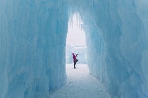 Plan your visit to the MN ice castles image of a caucasian woman standing in an ice castle in the entrance way and taking pictures on a cold winter day.