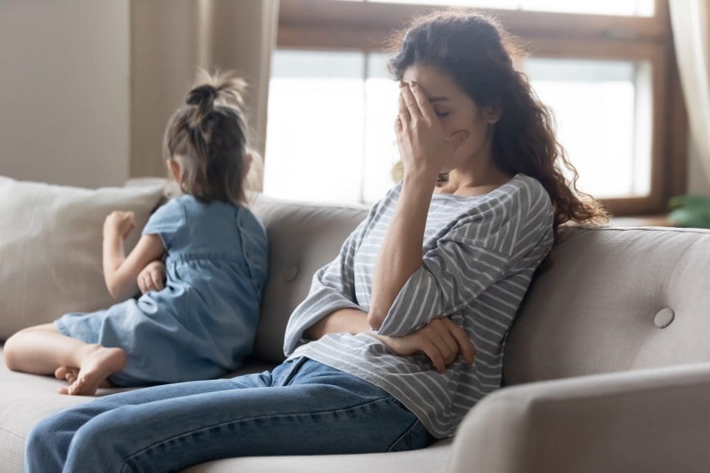 stepmom guilt - Unhappy young mother touching forehead, feeling tired of bad daughter's behavior at home. Offended little child girl sitting on different side on couch, ignoring sad frustrated mother in living room.