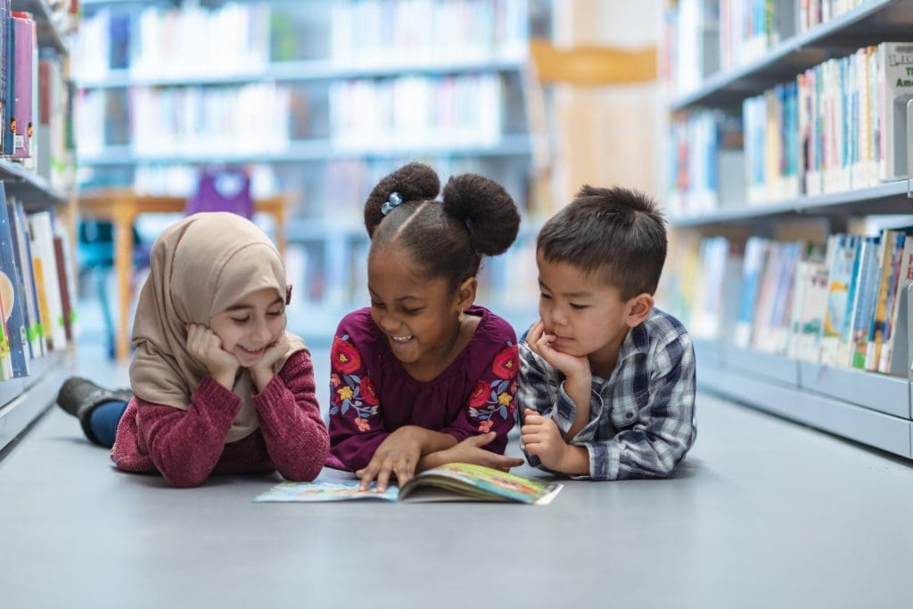 Three (3) kids who are friends are laying on the floor in between bookshelves in the library. They are reading a book together. They are all smiling and enjoying their afternoon