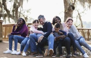 3 Transition Day Tips for Stepparents -A multi-ethnic blended family playing in the park together on a sunny day. They are sitting together on a deck. The African-American mother and Caucasian father have mixed race twin boys, almost 3 years old. Their daughters and step-daughters are 11 to 15 years old. The mother and girls are smiling and looking, looking at the camera. -