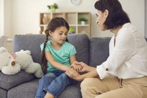 Young mother sitting on sofa with daughter, holding her hands and talking to her seriously at home with room interior at background. Resources for speaking to children about school shootings