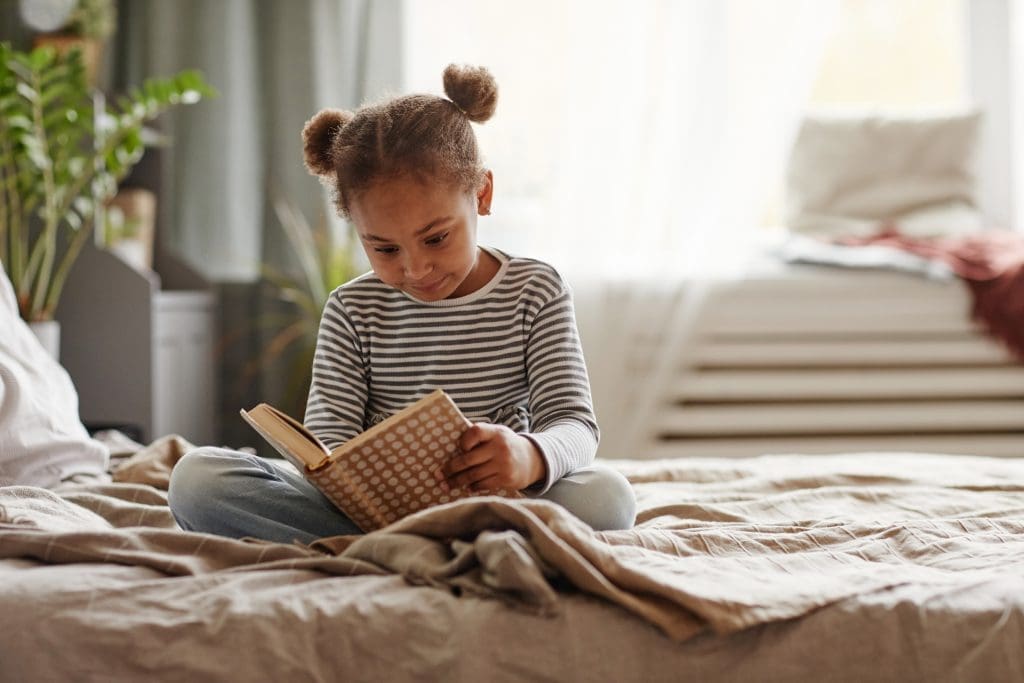 Summer Checklist - Full length portrait of cute African-American girl reading book while sitting on bed in cozy interior, copy space